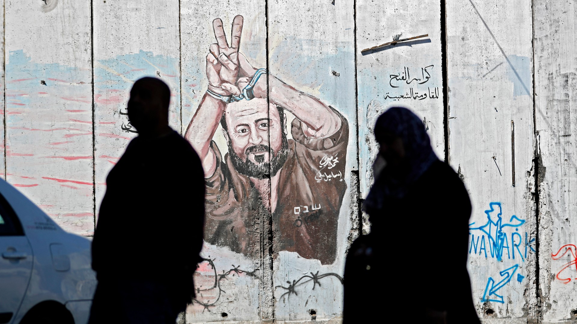 Palestinians walk past graffiti depicting Palestinian prisoner Marwan Barghouti in the occupied West Bank city of Abu Dis on 13 March 2018 (Thomas Coex/AFP)